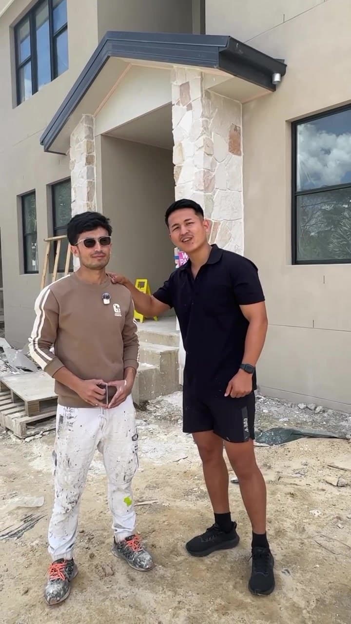 Mahdi, in a black polo shirt, stands beside Murtaza outside a new two-storey house on a building site. Murtaza wears paint-flecked white work trousers.